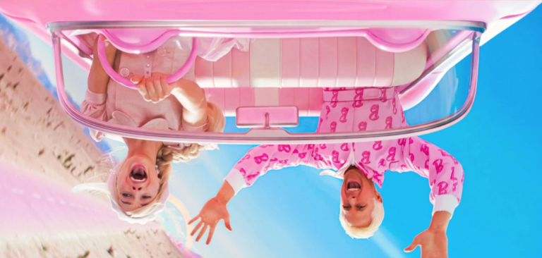 Cena do filme Barbie: Ela está dirigindo e no banco de trás está o Ken, o carro está de cabeça para baixo. e os dois estão com a boca aberta e assustados. Além de estarem vestido de rosa.