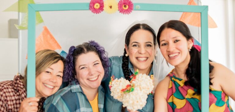 Texto Fechamento do mês: A imagem mostra quatro mulheres sorridentes participando de uma celebração festiva. Elas estão posando juntas dentro de uma moldura decorativa azul-clara que emoldura seus rostos. Da esquerda para a direita: A primeira mulher tem cabelo loiro, veste uma camisa xadrez marrom e segura a moldura com uma das mãos. Ela sorri amplamente. A segunda mulher tem cabelo cacheado tingido de roxo, preso em duas marias-chiquinhas, e está usando uma camisa xadrez azul. Ela também está sorrindo. A terceira mulher tem cabelo escuro preso em uma trança, está usando uma camisa xadrez azul e um véu de noiva. Ela segura um buquê de pipoca decorado com pequenas flores coloridas e também sorri amplamente. A quarta mulher tem cabelo escuro preso em um rabo de cavalo e veste um vestido colorido com estampas vibrantes em amarelo, verde e rosa. Ela também está sorrindo e segurando a moldura com uma das mãos. Ao fundo, há bandeirinhas coloridas típicas de festas juninas penduradas, reforçando o clima festivo da imagem. As mulheres estão com maquiagem que imita sardas no rosto, um elemento comum nas festas juninas brasileiras. A imagem transmite um sentimento de alegria, união e celebração.