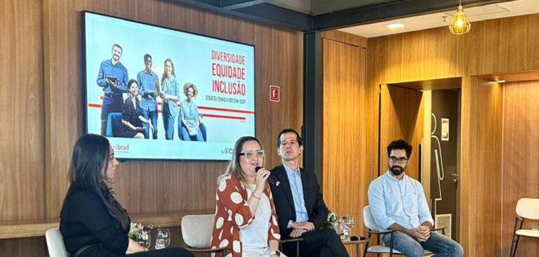 A imagem mostra uma cena de um painel de discussão ou conferência sobre diversidade, equidade e inclusão. Quatro pessoas estão sentadas em cadeiras, dispostas em uma linha, com um grande monitor ou tela de TV atrás deles, exibindo o título "Diversidade, Equidade, Inclusão" e a frase "O que isso tem a ver com ESG?", além de fotos de diversas pessoas ao fundo. Da esquerda para a direita: A primeira pessoa é uma mulher com cabelos longos e escuros, vestindo uma blusa preta. Ela está olhando para a segunda pessoa. A segunda pessoa é uma mulher com cabelos loiros, vestindo uma blusa branca e um casaco marrom com estampas de bolinhas brancas. Ela está segurando um microfone e falando. A terceira pessoa é um homem com cabelo curto e escuro, vestindo um terno escuro e camisa clara. Ele está olhando para a frente com uma expressão neutra. A quarta pessoa é um homem com cabelo curto e barba, vestindo uma camisa azul clara. Ele está sentado com as mãos cruzadas e também olhando para a frente. O fundo da sala é decorado com painéis de madeira, e à direita, há uma porta com um símbolo que indica um banheiro. A iluminação é suave e há luminárias pendentes visíveis no teto. A configuração sugere um ambiente formal de discussão, onde os participantes estão engajados em um diálogo sobre temas importantes relacionados à inclusão e diversidade no contexto empresarial e de responsabilidade social.