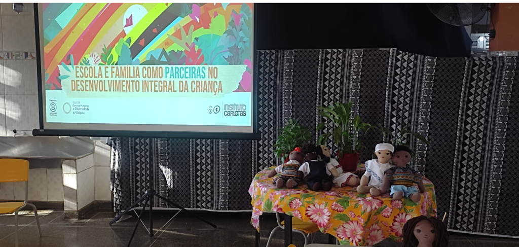 A imagem mostra um ambiente interno preparado para uma apresentação ou evento educacional. À esquerda, há uma tela de projeção grande com uma apresentação exibida. Na tela, lê-se o título "Escola e família como parceiras no desenvolvimento integral da criança", com ilustrações coloridas de elementos naturais como plantas e flores ao fundo. A apresentação é do Instituto Carlota, e há logos de certificações e edições na parte inferior esquerda da tela. À direita da imagem, há uma mesa coberta com uma toalha floral colorida, predominantemente em amarelo com flores rosas e brancas. Em cima da mesa, há várias bonecas de pano de diferentes etnias e vestimentas tradicionais, além de alguns vasos de plantas, sugerindo um ambiente acolhedor e diversificado. O fundo é decorado com um tecido preto e branco com padrões geométricos. À esquerda, há também uma cadeira amarela e uma pia de inox que parece fazer parte do local. A imagem transmite um ambiente de aprendizado e inclusão, destacando a parceria entre escola e família no desenvolvimento das crianças, com um enfoque em diversidade cultural e acolhimento.