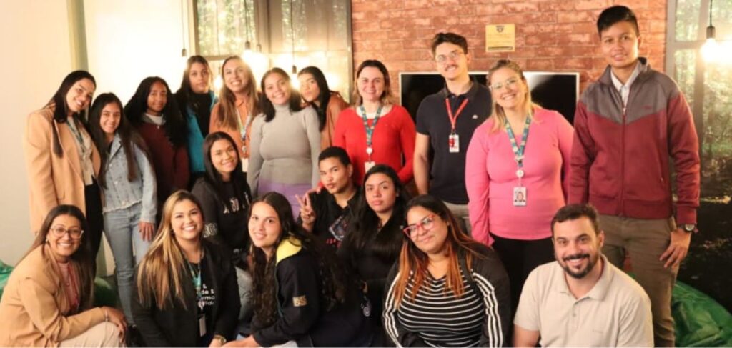 A imagem mostra um grupo de aproximadamente vinte pessoas, todas sorrindo, posando para uma foto de grupo em um ambiente interno que parece ser um espaço de convivência ou uma sala de reunião. As pessoas estão distribuídas em duas fileiras: uma em pé e outra agachada ou sentada na frente. Na fileira de trás, da esquerda para a direita: Uma mulher de cabelo escuro e liso, vestindo um blazer bege. Uma mulher de cabelo longo e escuro, vestindo uma jaqueta jeans. Uma mulher de cabelo escuro e cacheado, vestindo uma blusa vermelha. Uma mulher de cabelo escuro, vestindo um suéter laranja. Uma mulher de cabelo castanho, vestindo uma blusa cinza de manga longa. Uma mulher de cabelo preto e longo, parcialmente escondida atrás de outras pessoas. Uma mulher de cabelo castanho claro, vestindo uma blusa vermelha com crachá. Um homem de cabelo curto, vestindo uma camisa polo preta com crachá. Uma mulher de cabelo loiro, vestindo uma blusa rosa e óculos. Um homem de cabelo curto, vestindo uma jaqueta cinza e bordô. Na fileira da frente, da esquerda para a direita: Uma mulher de cabelo longo e liso, usando óculos e uma camisa bege. Uma mulher de cabelo loiro, vestindo uma blusa preta com crachá. Uma mulher de cabelo longo e escuro, vestindo uma blusa preta. Um homem de cabelo curto e escuro, fazendo um sinal de paz com os dedos. Uma mulher de cabelo longo e escuro, vestindo uma blusa preta com detalhes amarelos. Uma mulher de cabelo longo e com mechas loiras, vestindo uma blusa listrada preta e branca. Um homem de cabelo curto e barba, vestindo uma camisa polo cinza. O fundo da imagem mostra uma parede de tijolos aparentes e janelas com luz natural entrando. Há também uma televisão ou tela grande na parede e algumas luzes pendentes. A atmosfera é acolhedora e amigável, sugerindo que o grupo está participando de um evento ou reunião relacionada a responsabilidade social e ESG (ambiental, social e governança).