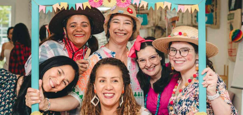 festa junina A imagem mostra seis mulheres sorrindo e posando juntas em uma festa temática, provavelmente uma festa junina. Elas estão vestindo roupas típicas para a ocasião, com chapéus de palha decorados, roupas coloridas e alguns acessórios festivos, como lenços e laços no cabelo. As mulheres estão segurando uma moldura azul turquesa decorada com pequenas bandeirinhas coloridas na parte superior, que adiciona um toque festivo à cena. O ambiente ao fundo sugere que estão em um espaço interno decorado para a festa, com mais bandeirinhas e outros elementos típicos ao fundo. Todas estão felizes e parecem estar aproveitando o momento juntas.