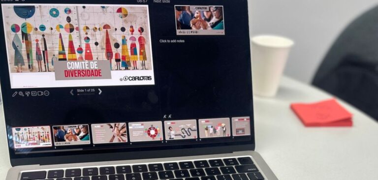 A imagem mostra um laptop aberto em cima de uma mesa, exibindo uma apresentação de slides em uma tela de software de apresentação, como o PowerPoint ou Google Slides. O slide que está sendo mostrado na tela é intitulado "Comitê de Diversidade", com o logo "Carlotas" visível no canto inferior direito. O slide inclui uma imagem colorida e abstrata, com formas geométricas e figuras estilizadas de pessoas, refletindo o tema da diversidade. Na parte inferior da tela do laptop, há uma visualização dos slides em miniatura, sugerindo que a apresentação possui 25 slides no total, com o slide atual sendo o primeiro. Ao lado da tela do laptop, há um copo descartável branco e um bloco de notas vermelho, que estão sobre a mesa cinza. O ambiente sugere uma reunião ou workshop onde o tema da diversidade está sendo discutido, possivelmente em um ambiente corporativo ou educacional. (CAPA Empresas - Agosto Fechamento)
