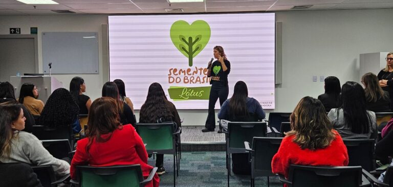 A imagem mostra uma apresentação em um auditório. Uma mulher está em pé na frente de uma tela grande, segurando um microfone e falando para o público. Na tela, há um logotipo verde em forma de coração com um broto de planta no centro, acompanhado do texto "Semente do Brasil" e "Lótus". O público, composto majoritariamente por mulheres, está sentado em cadeiras, prestando atenção na palestrante. As cadeiras estão dispostas em fileiras, e as pessoas estão voltadas para a frente. Algumas das mulheres estão usando roupas coloridas, incluindo uma mulher em um casaco vermelho. O ambiente é moderno e bem iluminado, com paredes claras e uma atmosfera profissional. Ao fundo, à esquerda da tela, há uma mesa com um computador e um microfone, e ao lado direito, uma mulher de pé observa a apresentação. A sala parece ser um espaço de conferências ou uma sala de aula equipada para apresentações multimídia.