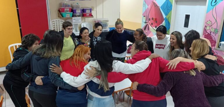 A imagem mostra um grupo de mulheres em uma sala, formando um círculo e abraçadas pelos ombros. Todas parecem estar participando de uma atividade coletiva, exibindo expressões de alegria e camaradagem. A sala está decorada de maneira acolhedora e colorida. Ao fundo, há armários com caixas plásticas de armazenamento, algumas cadeiras empilhadas e uma parede colorida com murais artísticos que incluem desenhos de flores e formas abstratas. À esquerda, uma parede amarela e vermelha adiciona ainda mais cor ao ambiente. As mulheres estão vestidas de forma casual, com uma variedade de roupas coloridas. Algumas delas usam jaquetas, enquanto outras vestem camisetas. A atmosfera da imagem é descontraída e amigável, sugerindo um momento de união e apoio mútuo entre as participantes.