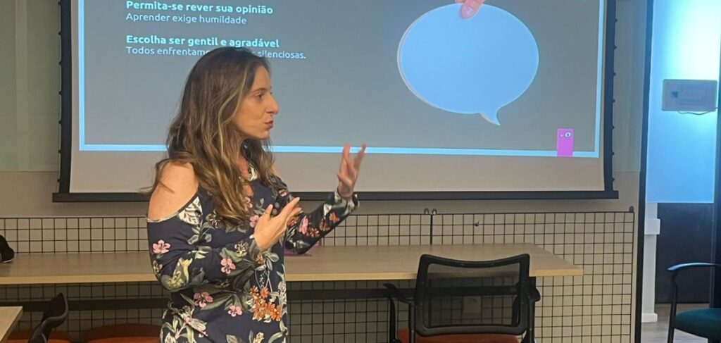 Julho: A imagem mostra uma mulher apresentando em frente a uma tela de projeção em uma sala de conferências ou aula. Ela está de perfil, olhando para a tela, e gesticula com as mãos enquanto fala. A mulher tem cabelo castanho claro, longo e ondulado, e está vestindo uma blusa de mangas compridas com estampa floral. Na tela de projeção, há texto e gráficos, incluindo uma imagem de um balão de fala azul. O texto visível na tela diz: "Permita-se rever sua opinião" "Aprender exige humildade" "Escolha ser gentil e agradável" "Todos enfrentamos batalhas silenciosas." A sala é equipada com mesas e cadeiras, e a decoração é simples e funcional, com uma parede de azulejos quadrados ao fundo. A iluminação é clara e uniforme, proporcionando um ambiente propício para apresentações e discussões. A atmosfera da imagem sugere um ambiente de aprendizado e compartilhamento de conhecimentos, com a palestrante engajada em transmitir informações importantes ao seu público.