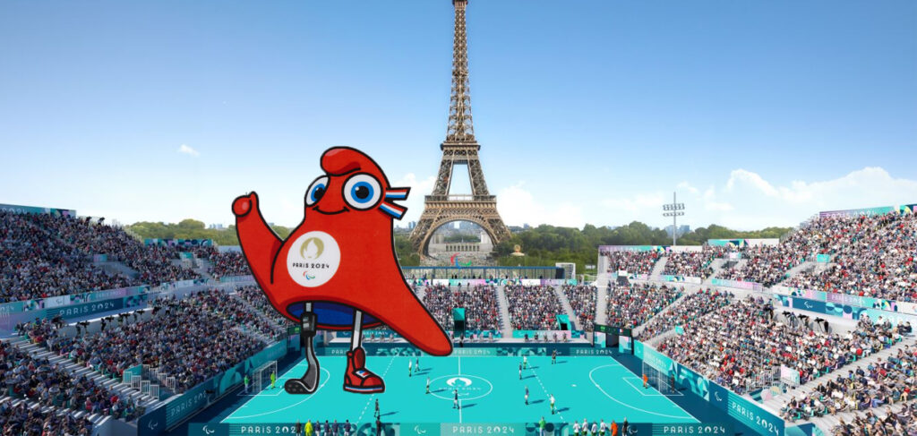 A imagem mostra um estádio lotado em Paris, com a icônica Torre Eiffel ao fundo. No centro da imagem, aparece a mascote oficial dos Jogos Paralímpicos de Paris 2024, com uma expressão alegre e um olho grande. A mascote tem uma prótese na perna esquerda, simbolizando a inclusão e a diversidade dos atletas paralímpicos.