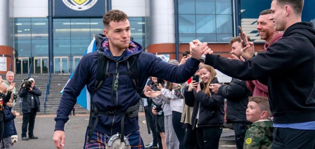 Nesta imagem, vemos um homem jovem, vestido com roupas esportivas, incluindo uma jaqueta azul marinho e uma saia de tartan, o que sugere um toque cultural escocês. Ele está cercado por um grupo de pessoas que o aplaudem e interagem com ele, muitas delas sorrindo e tirando fotos com seus celulares. O homem parece estar cumprimentando alguém à sua frente com um aperto de mão, enquanto mantém uma expressão confiante e orgulhosa. Ele carrega uma mochila, possivelmente indicando que ele está em uma caminhada ou desafio físico. Ao fundo, podemos ver o que parece ser um prédio grande, possivelmente um estádio ou uma arena, com pessoas ao redor, o que sugere que o evento ou encontro pode estar acontecendo em um local público