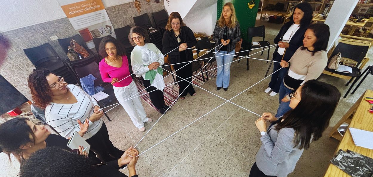 A imagem mostra um grupo de mulheres participando de uma dinâmica em um ambiente interno. Elas estão em pé, formando um círculo e segurando fios de lã, que se cruzam entre elas, criando uma teia no centro. A atividade parece ser colaborativa e simbólica, talvez relacionada à construção de conexões e interações entre as participantes. O local aparenta ser uma sala multiuso, com cadeiras empilhadas ao fundo. Há um cartaz na parede com o texto "Nosso objetivo é diminuir distâncias", sugerindo que a atividade tem um foco em integração ou trabalho em equipe. O ambiente é iluminado e há uma mesa ao lado com alguns materiais de papelaria. A dinâmica parece ser uma forma de reforçar o conceito de interconexão entre as participantes, incentivando o trabalho em grupo. 