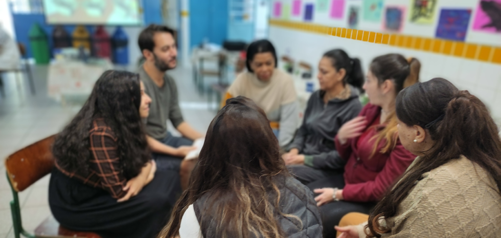 Um grupo de pessoas sentadas em círculo em um ambiente escolar, participando de uma atividade ou conversa coletiva. O local apresenta paredes decoradas com desenhos coloridos e lixeiras ao fundo. As pessoas estão engajadas, demonstrando interação e troca de ideias, sugerindo um momento de aprendizado ou colaboração em grupo.