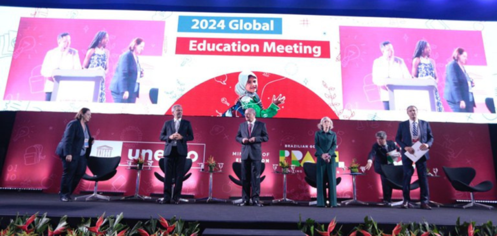 Imagem da "Reunião Global de Educação 2024". Seis pessoas estão no palco, com um telão ao fundo exibindo o título do evento e a imagem de uma jovem sorrindo. Logos da UNESCO e do Ministério da Educação do Brasil são visíveis.