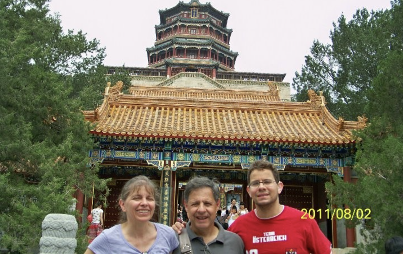 Três pessoas posando em frente a uma estrutura histórica chinesa com arquitetura tradicional, composta por telhados dourados ornamentados. A imagem está cercada por árvores, e há uma data marcada no canto inferior direito: "2011/08/02".