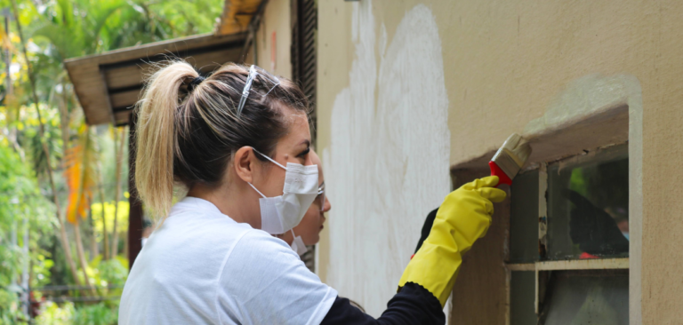 Mulher usando máscara facial e luvas amarelas, pintando a parte externa de uma janela em uma parede bege. A cena ocorre em um ambiente arborizado e reflete a colaboração em ações de voluntariado. A imagem está relacionada ao artigo sobre voluntariado empresarial.