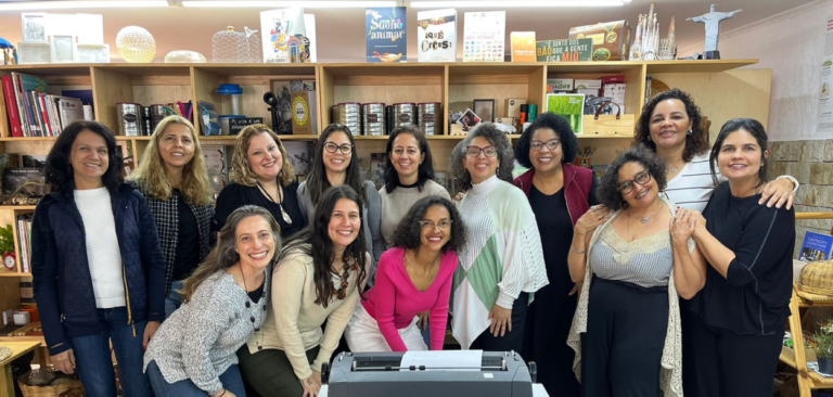 Foto de um grupo de gestoras e educadoras em um ambiente acolhedor, com estantes repletas de livros e objetos decorativos ao fundo. Todas estão sorrindo e posando juntas, transmitindo um clima de união e alegria. Algumas mulheres estão em pé e outras agachadas à frente, compondo uma formação descontraída