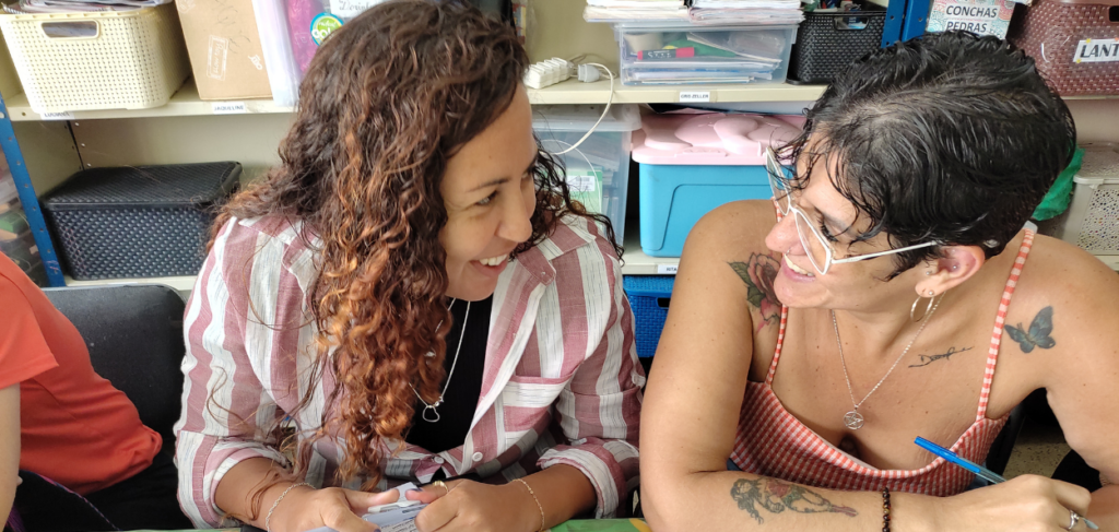 Duas Educadoras participantes do ExploreCarlotas se olham com cumplicidade e sorrisos