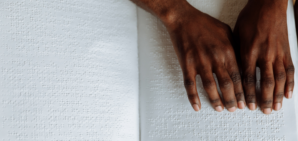 Mãos sobre um livro escrito em Braille. As duas mãos negras leêm o livro utilizando a leitura em Braille