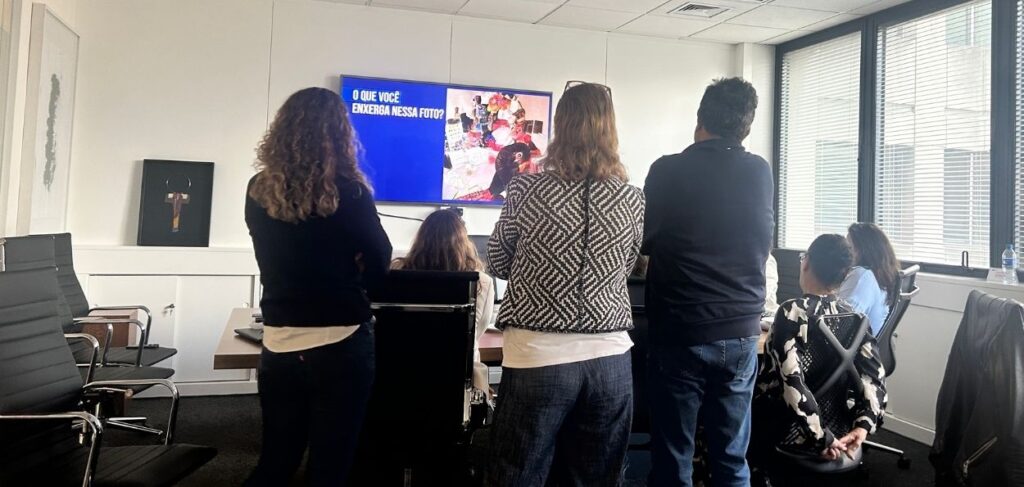 Colaboradores em sala de reunião observam imagem projetada com a pergunta “O que você enxerga nessa foto?”, durante atividade de sensibilização sobre diversidade e inclusão no ambiente corporativo.