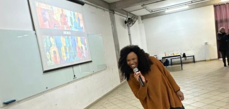 Adailza Barbosa, de Carlotas, sorri enquanto fala ao microfone em sala de aula durante o encerramento de uma atividade educativa. Ao fundo, uma projeção colorida exibe artes visuais com figuras humanas estilizadas.