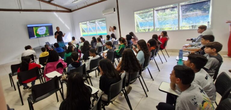 Crianças assistem a palestra sobre ESG em sala de aula, com educador explicando conceitos em uma apresentação na tela.